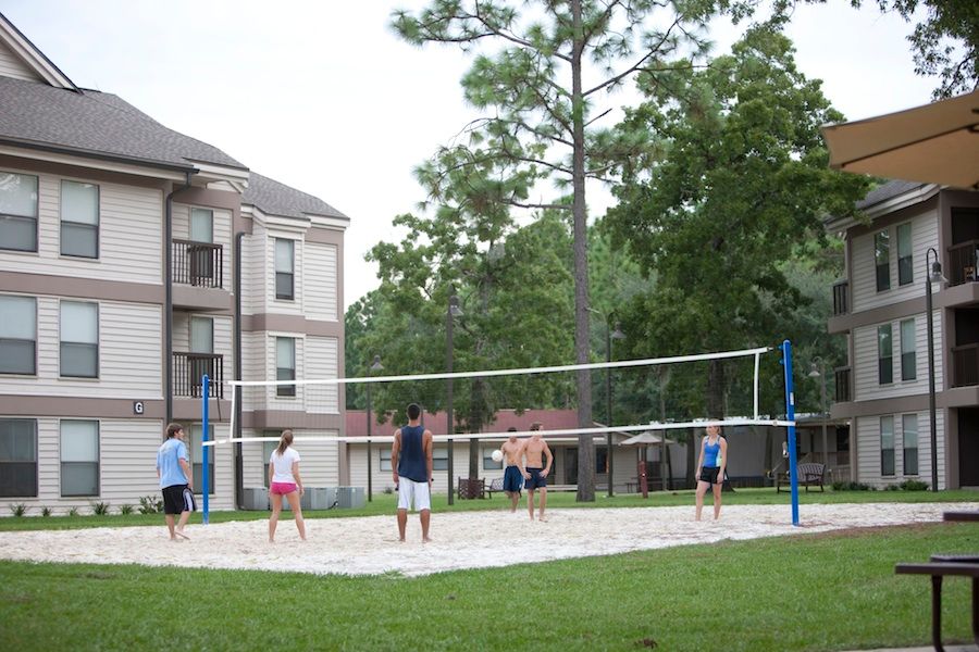 A group of six students plays volleyball in a sand pit within a grassy clearing between two off white and brown buildings; in the background are more buildings and a few tall pine trees.