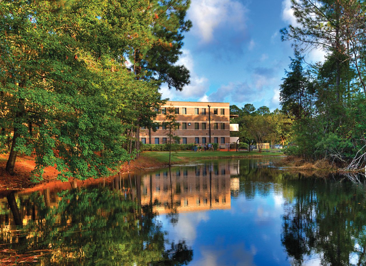 Three-story brick building with white trim sits on the far shore of a still lake with tall trees located along the shore