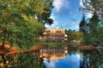 Three-story brick building with white trim sits on the far shore of a still lake with tall trees located along the shore