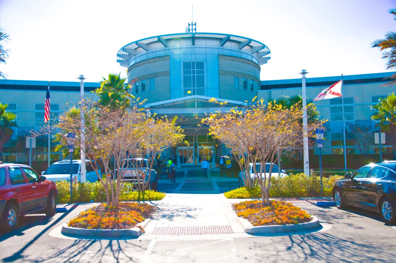 Two-story concrete and steel building with a wide and rounded front entrance sits at the end of a walkway lined with flowering trees.