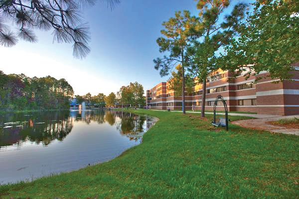 Three-story brick building with white trim sits along the right bank of a large lake surrounded by a wide grassy lawn; on the left bank of the lake is a dense patch of tall trees