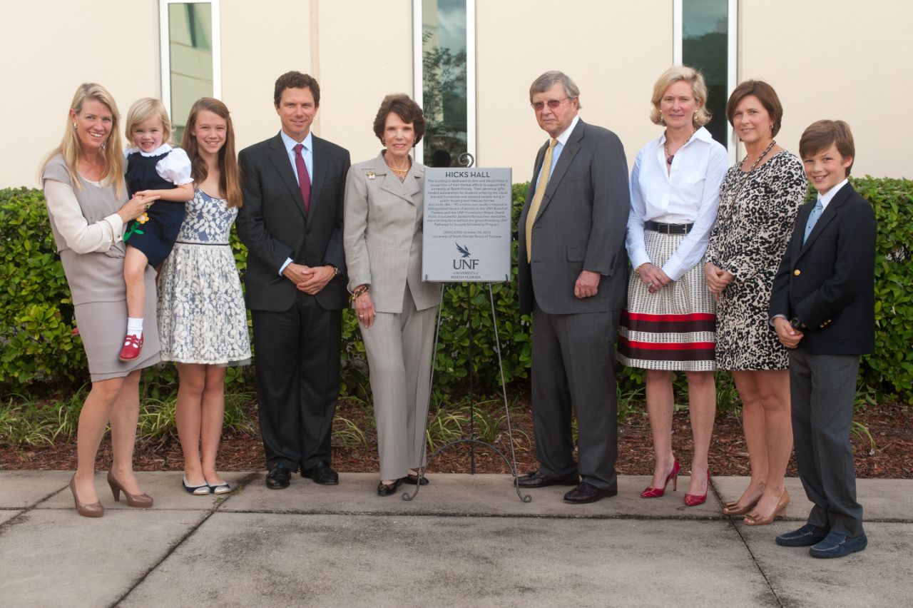 A multi-generational Caucasian family stands with a plaque on an easel outside in front of a two-story yellow stucco building on a cement patio with landscaping behind them. The plaque is titled “Hicks Hall” and bears the UNF logo.