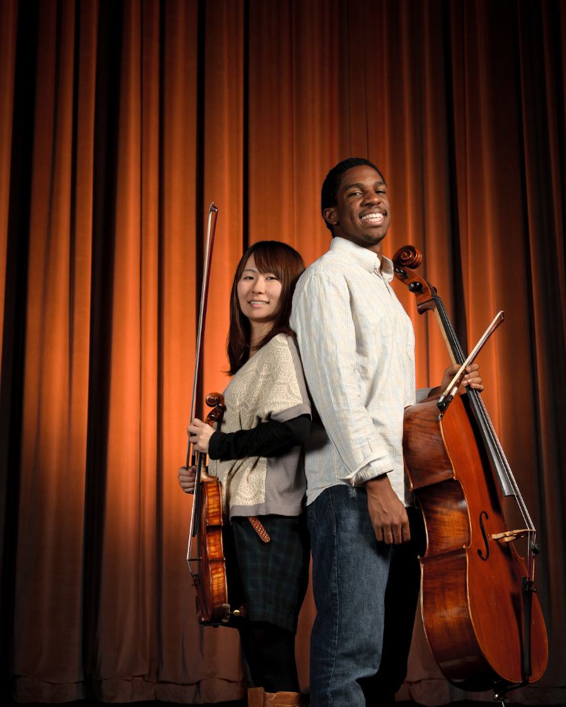 Two students stand back-to-back on a stage in front of an orange velvet curtain. The female student is holding a violin and the male student is holding a cello.