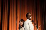 Two students stand back-to-back on a stage in front of an orange velvet curtain. The female student is holding a violin and the male student is holding a cello.