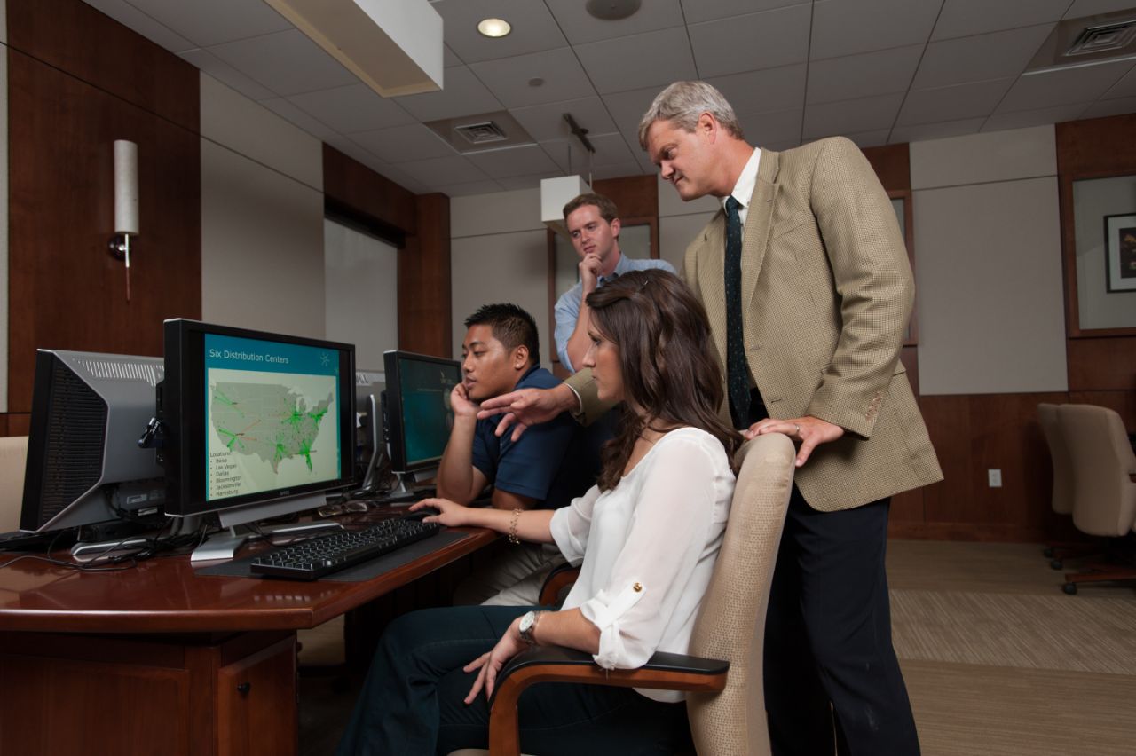 A professor points to a computer screen while three students look on in a computer lab.