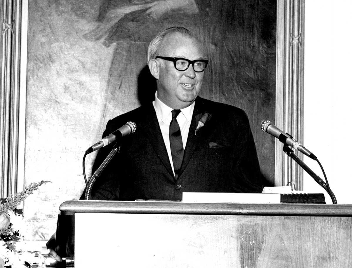 An older male stands at a podium with two microphones.