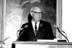 An older male stands at a podium with two microphones.