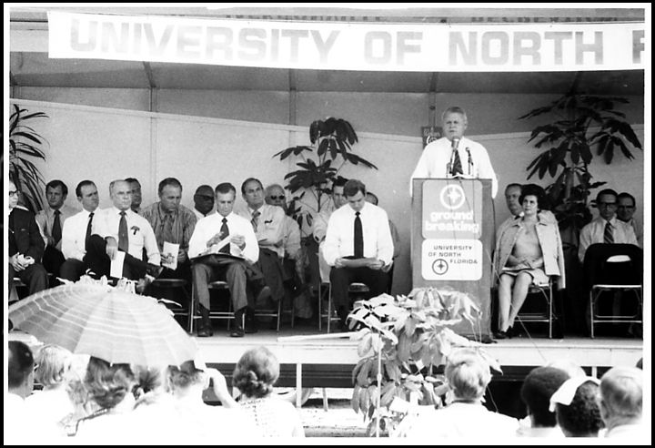 A group of people sitting on chairs on a stage before a large crowd listen to a man speaking from a podium.