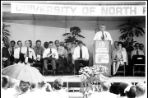 A group of people sitting on chairs on a stage before a large crowd listen to a man speaking from a podium.