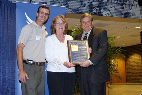 Two white males flank a white female holding a plaque while on a stage.