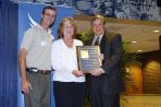 Two white males flank a white female holding a plaque while on a stage.