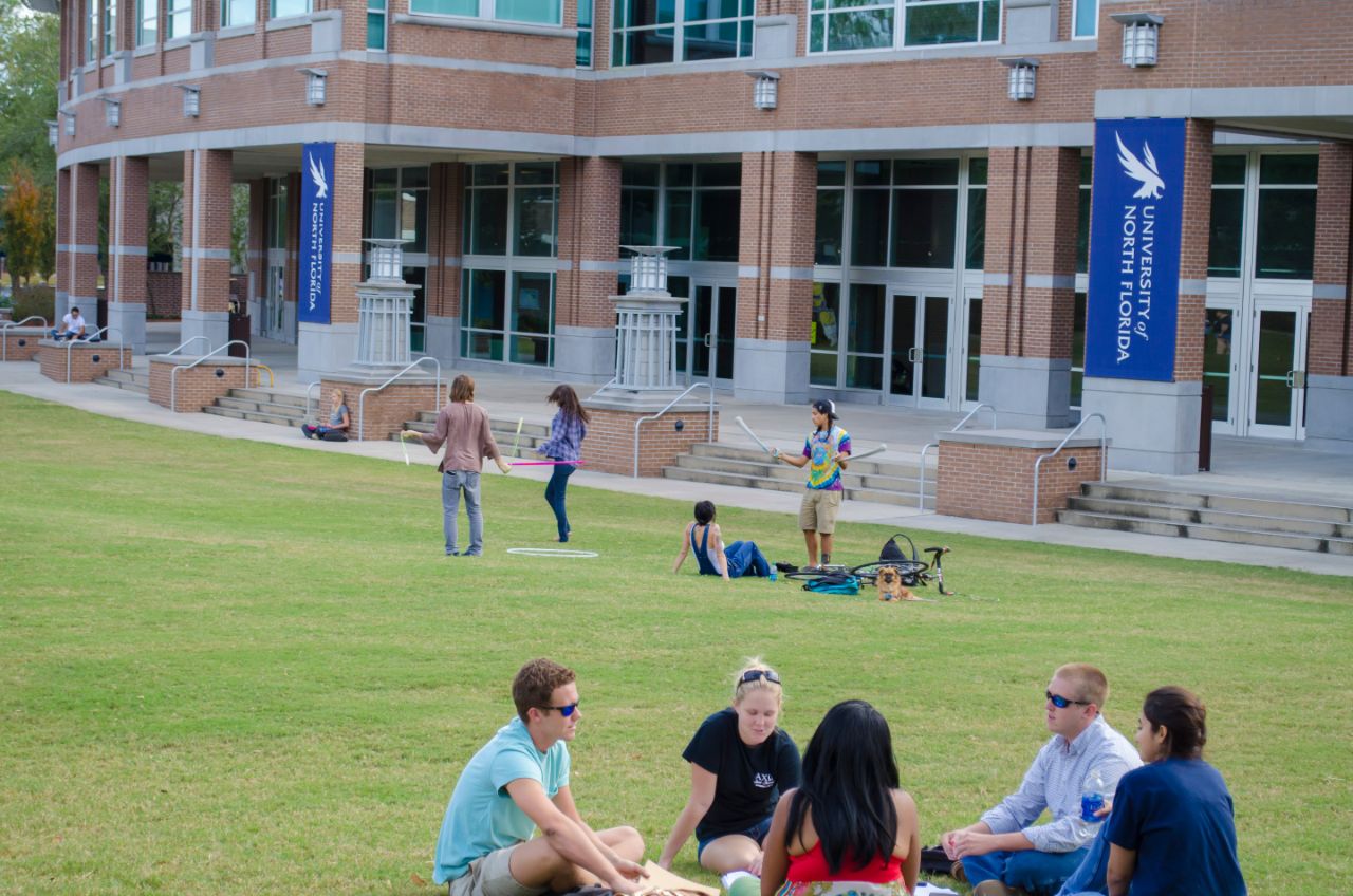 A group of five students sit in a circle in the foreground talking. Four other students are hulu hooping closer to the Fine Arts building.