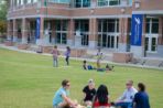 A group of five students sit in a circle in the foreground talking. Four other students are hulu hooping closer to the Fine Arts building.