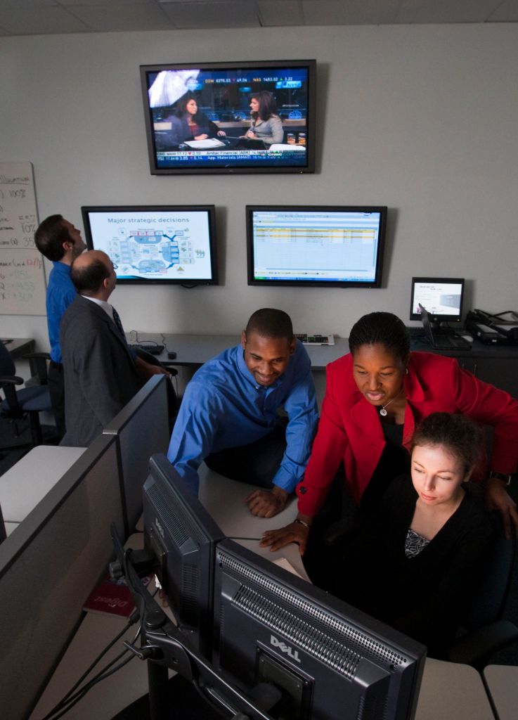 Two males stand in the background watching television while a female professor and two students group around a computer screen.