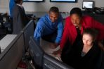 Two males stand in the background watching television while a female professor and two students group around a computer screen.