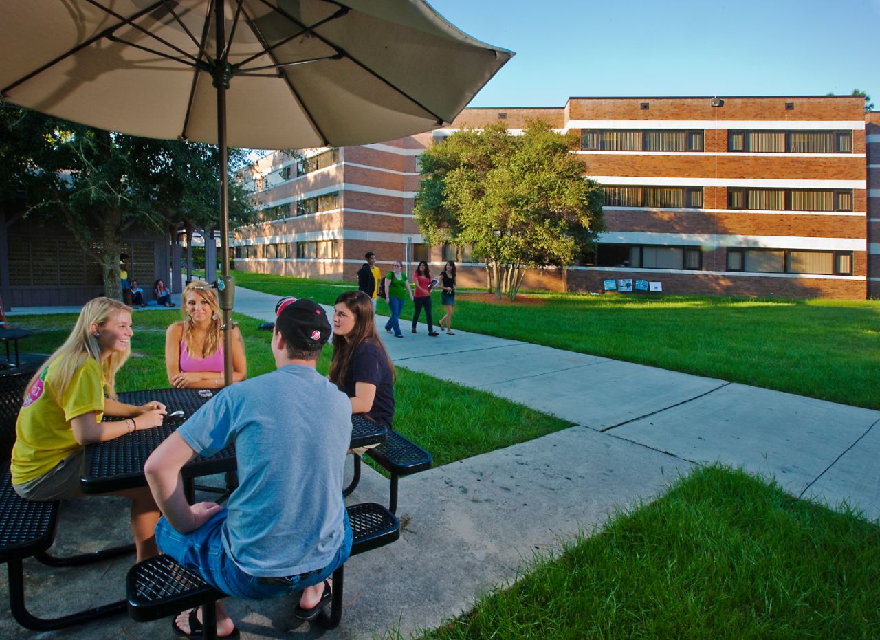 Three females and one male sitting at a picnic table under an umbrella talking. Three other females and a male are walking on the sidewalk behind the picnic table.