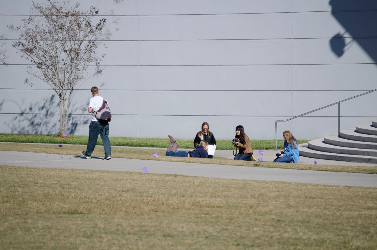 Four female students in front of Coxwell Amphitheater. Two of the students have cameras are taking photos of another female student laying in the grass. A male student is walking by on the side walk.