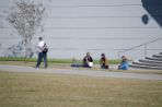 Four female students in front of Coxwell Amphitheater. Two of the students have cameras are taking photos of another female student laying in the grass. A male student is walking by on the side walk.