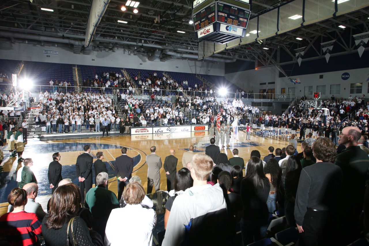 A large crowd of people in the arena stand while four memebers of the ROTC present the American and UNF flags.