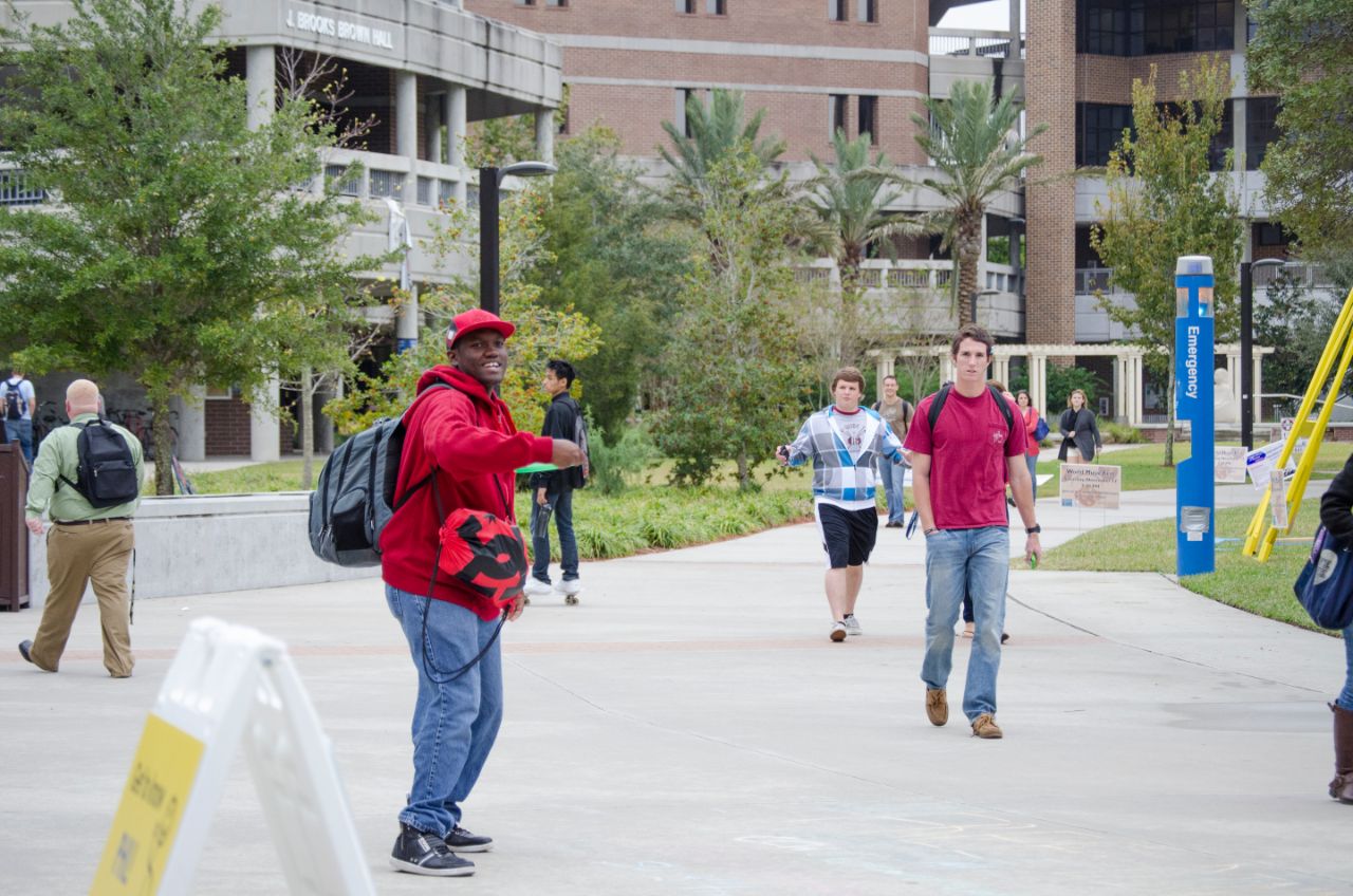 A male students throws a green frisbee towards the camera. More students are walking by on the sidewalk in front of the Brooks College of Health.