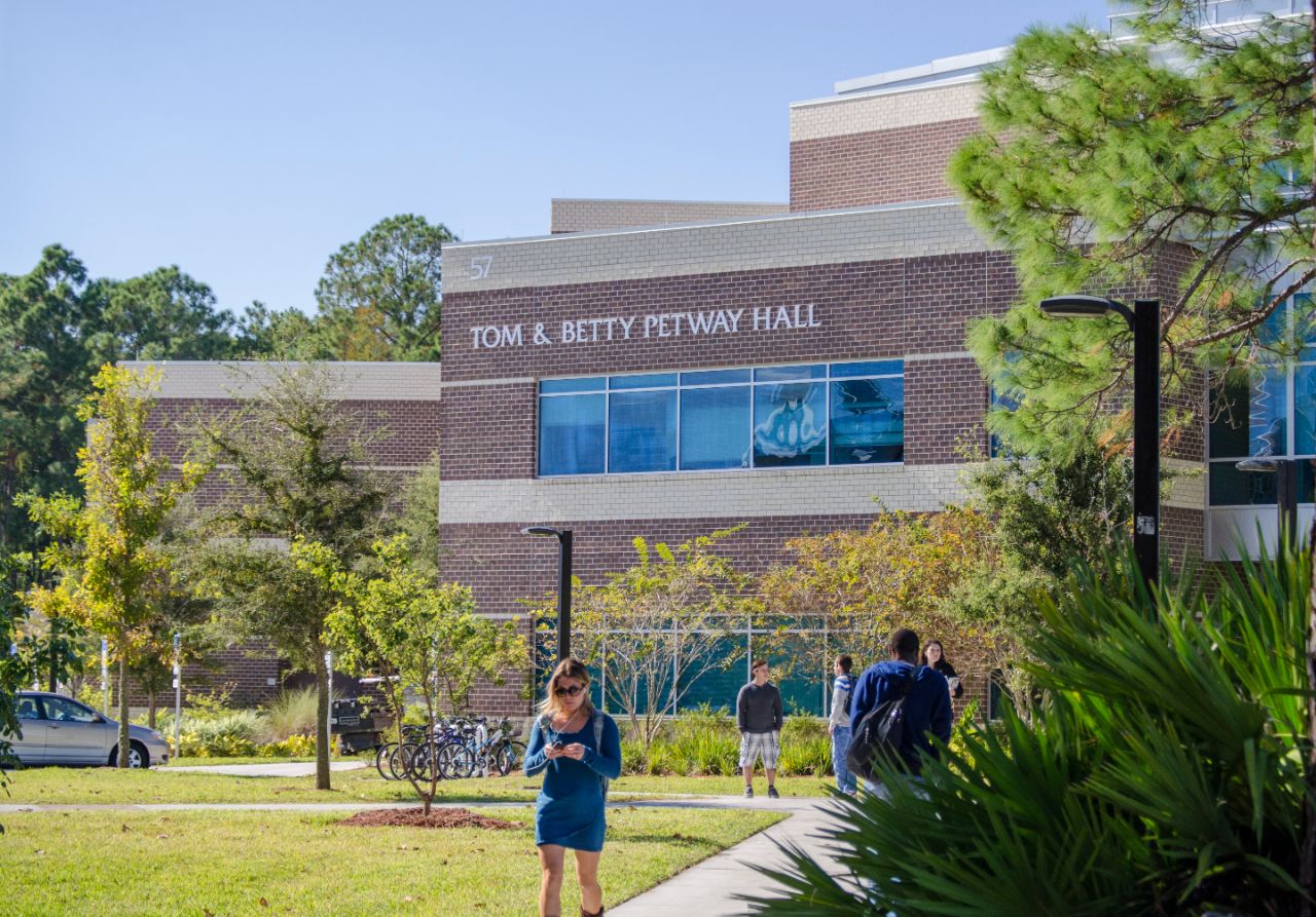 A female walking with her cell phone. A male with a backpack is walking towards Petway Hall.