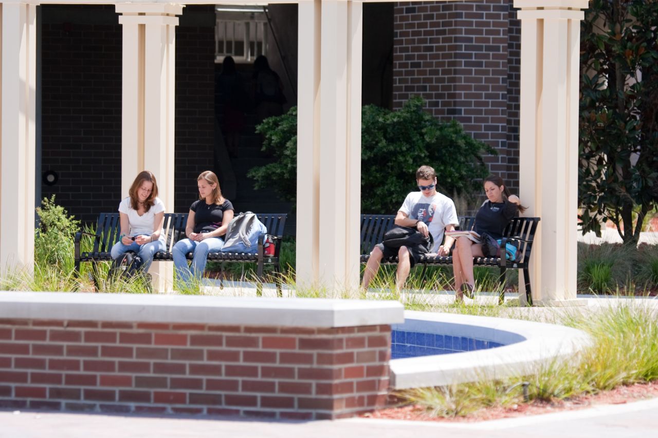 Three female students and one male student sit on benches at the Meditation Pond in front of the Brooks College of Health.