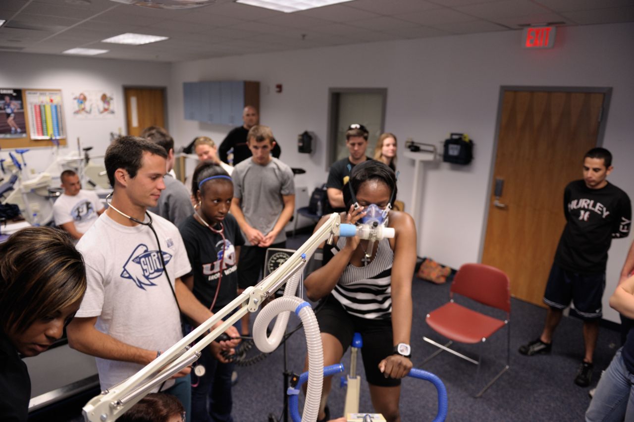 A female student is on an exercise bicycle with a respiration device on her head while other students monitor her progress.