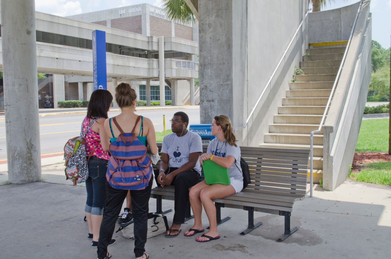 Two female students stand in front of a male and female student sitting on a bench talking.