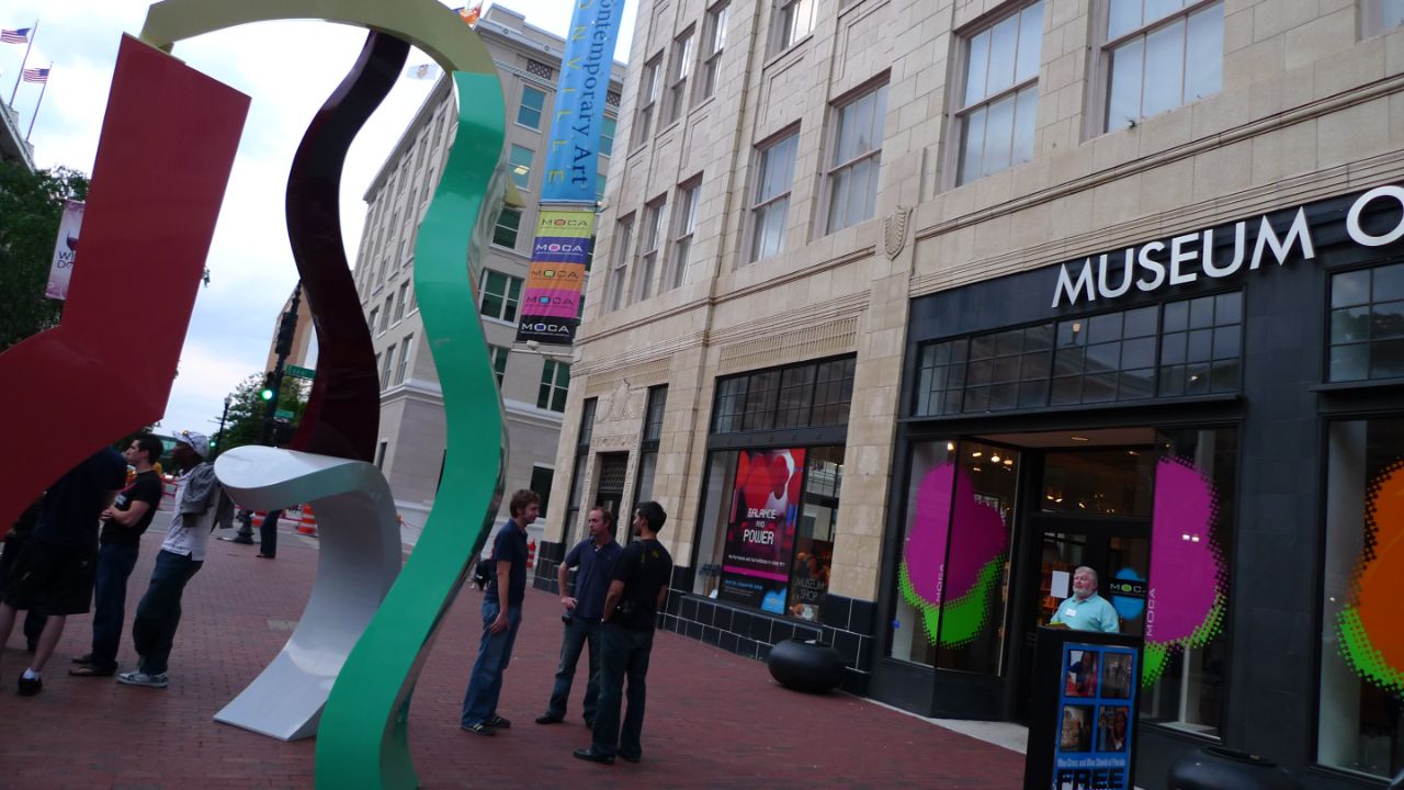 Three male students stand between the front door of the Museum of Contemporary Arts and a large colorful sculpture on the sidewalk.