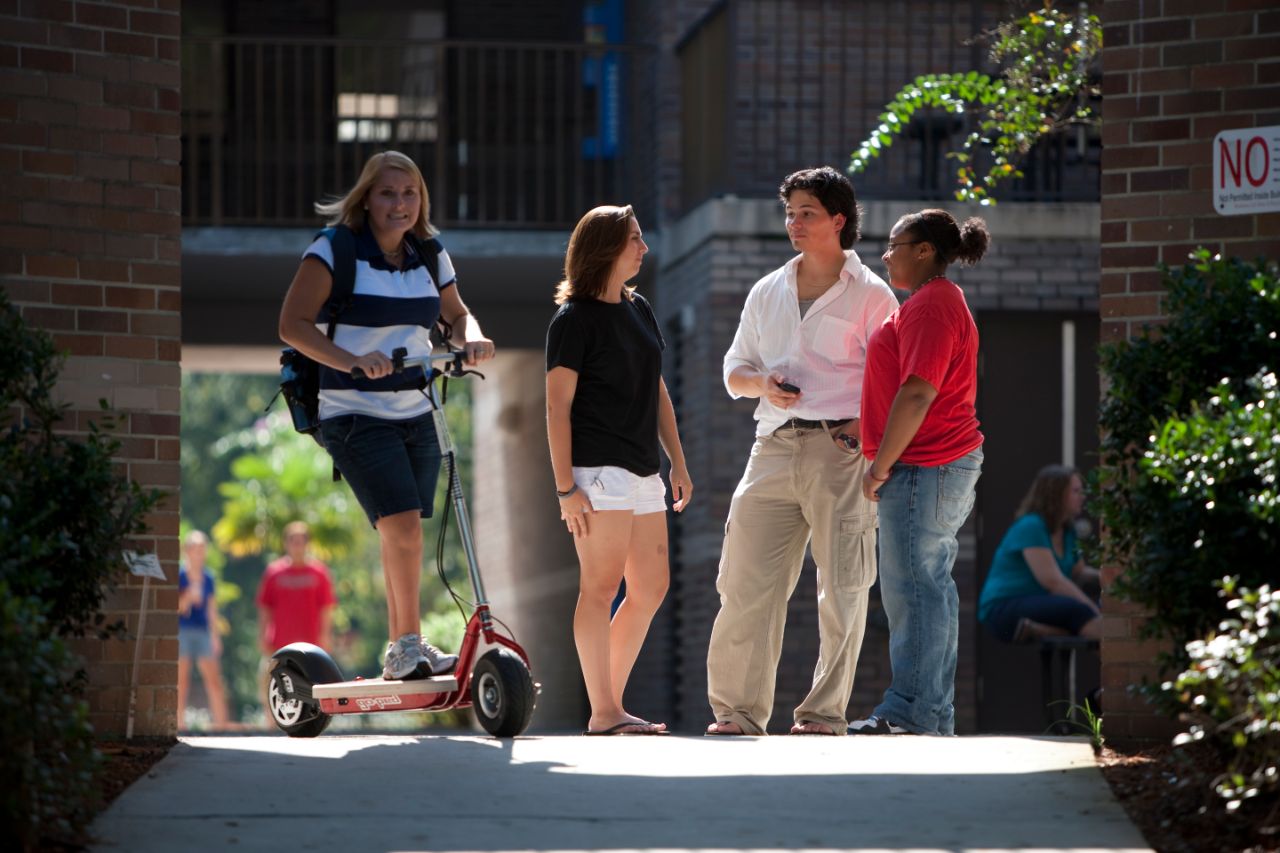 A female on an electric scooter with two females and one male talking behind her in a group.