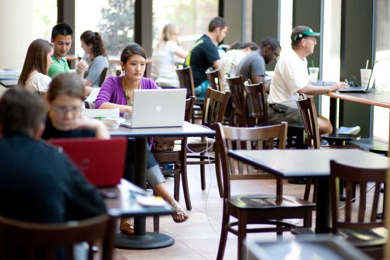 A lot of tables with the focus of a female working on her laptop.