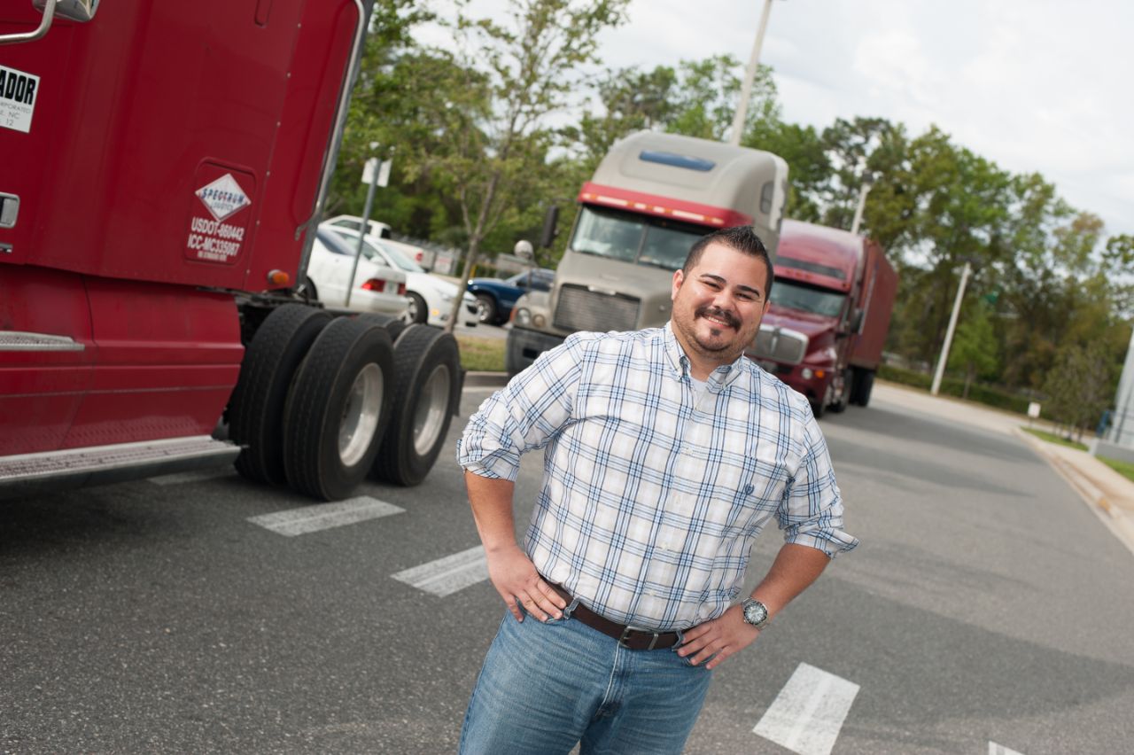 A Hispanic male stands in front of several 18-wheelers lined up behind him on a long driveway.