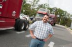 A Hispanic male stands in front of several 18-wheelers lined up behind him on a long driveway.