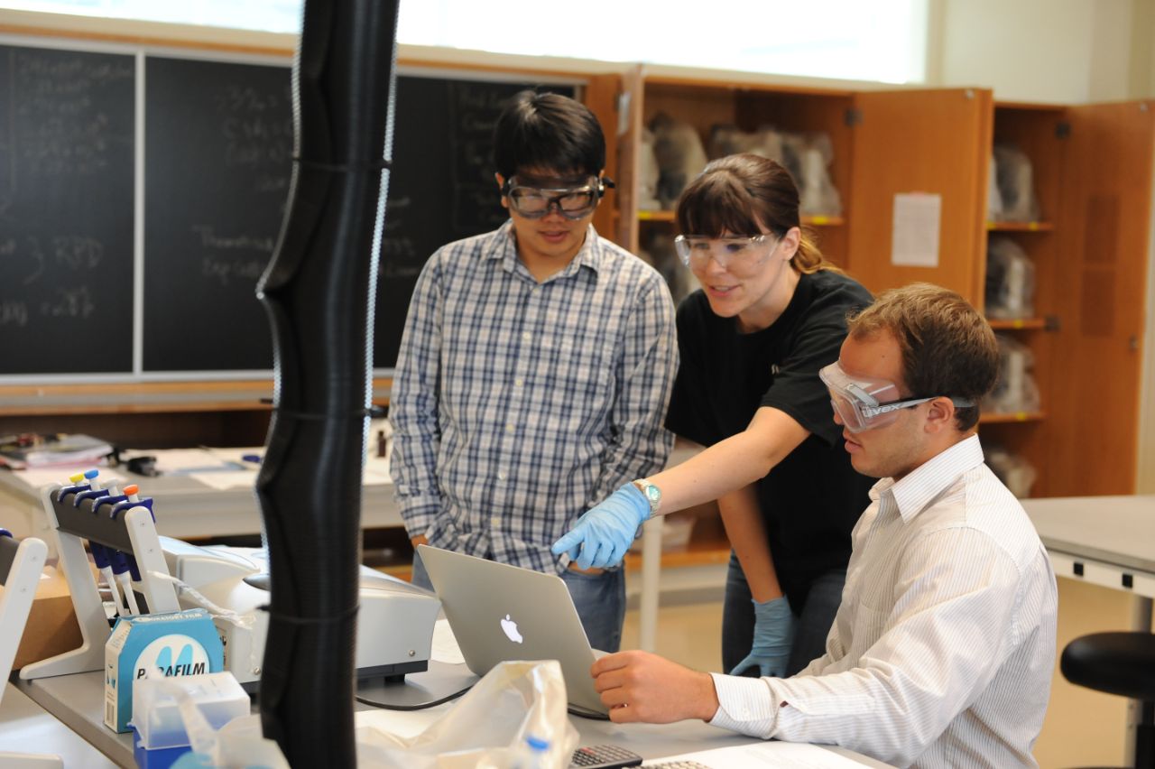 Two males students stand with a female professor at a bench lab table while wearing safety goggles in a laboratory.