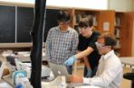 Two males students stand with a female professor at a bench lab table while wearing safety goggles in a laboratory.