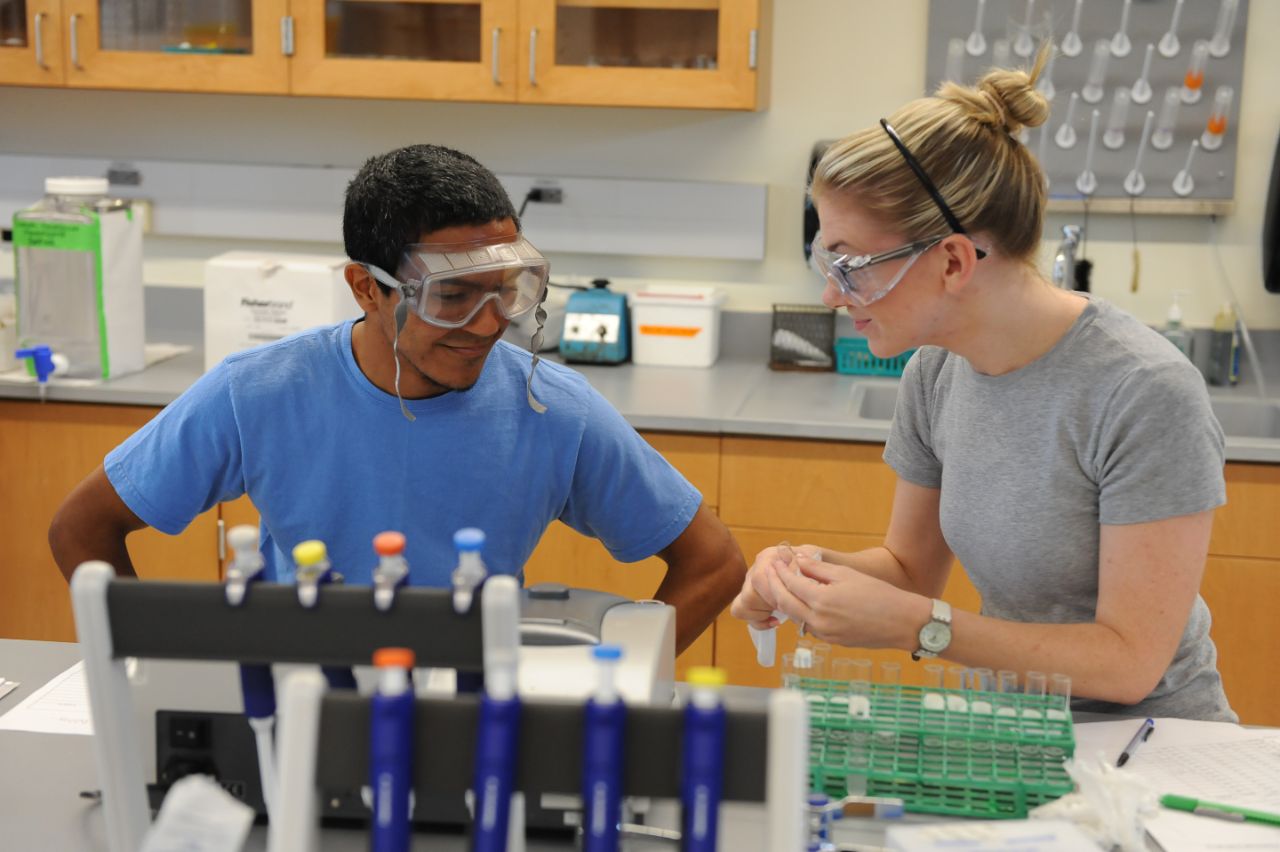 Two students handle science equipment at a bench table while wearing safety goggles in a laboratory.