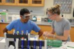 Two students handle science equipment at a bench table while wearing safety goggles in a laboratory.